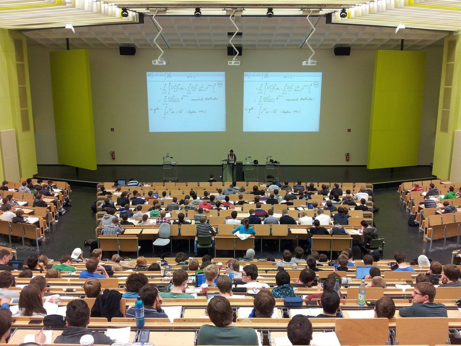 university lecture theatre showing students seated throughout theatre facing a lecturer with a large screen behind covered in equations