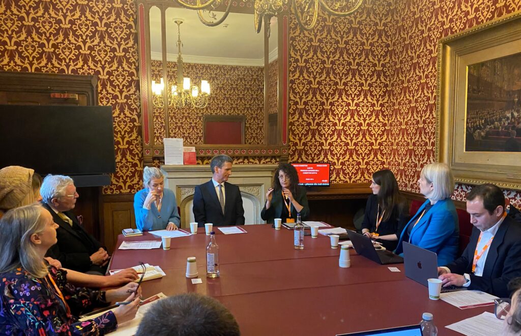 Large board table with several people seated around it, at head Chris, centre looks at the woman seated on his left who is gesturing with her hands.